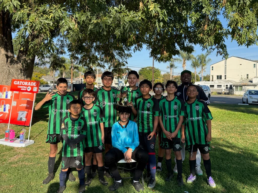 Phantom FC players in green and black stripes celebrating with a trophy under a shade tree